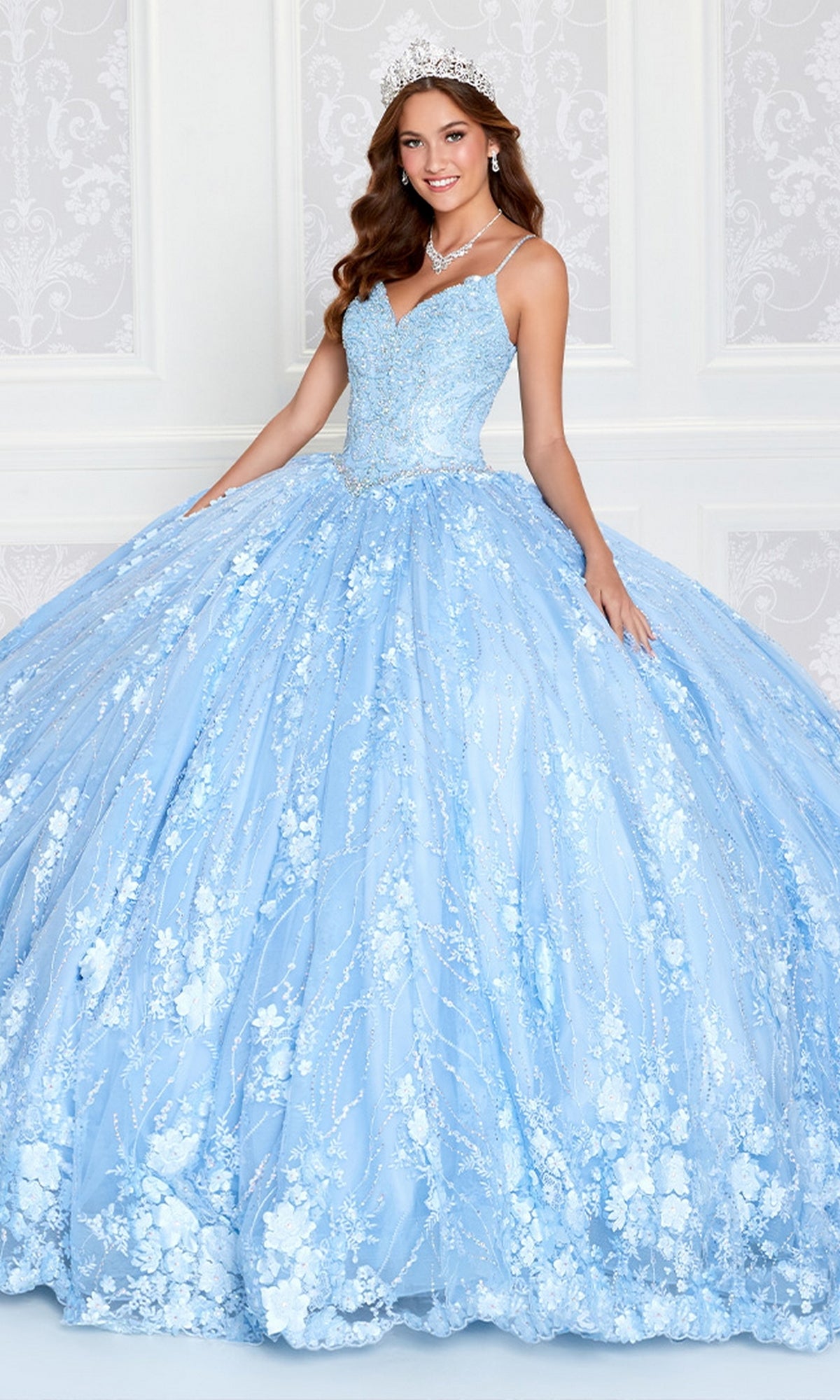 A woman in a sparkling light blue Princesa by Ariana Vara Lace Quince Dress PR12268 and tiara stands before a white patterned wall, smiling with her hands resting on the dress's voluminous skirt.